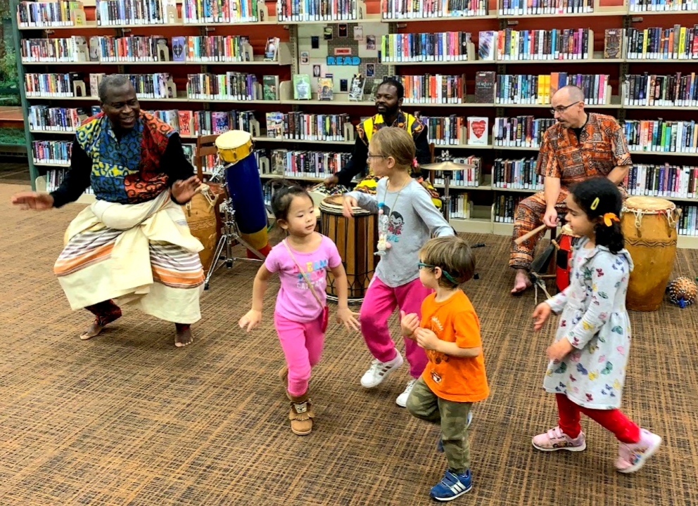 Etienne dancing with small children at a library with musicians Yaw, and Greg playing drums. 