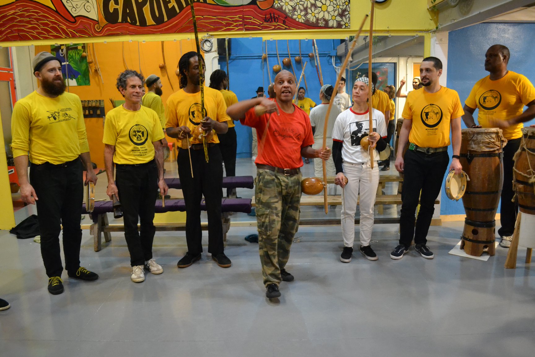 Silvio holding the Berimbau and teaching in a roda (capoeira cypher). They are in a brightly colored studio with many Berimbaus on the wall. 