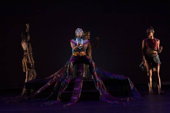 Rujeko Dumbutshna cups her hands upwards while wearing a magnificently colorful African textile skirt that sweeps down and across the pedestal on which she sits. 