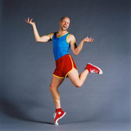 Bald man in bright athletic clothing humorously balances on one toe. 