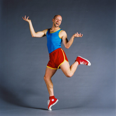 Bald man in bright athletic clothing humorously balances on one toe. 