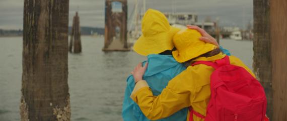 two in colorful raincoats and rain hats lean on each other on a dock 