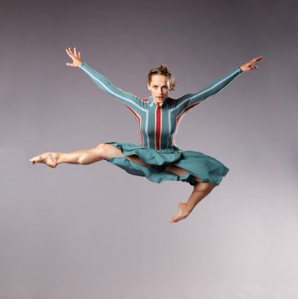 Muscular lady in striped dress jumps high with her arms out to her sides, and one foot tucked underneath. 