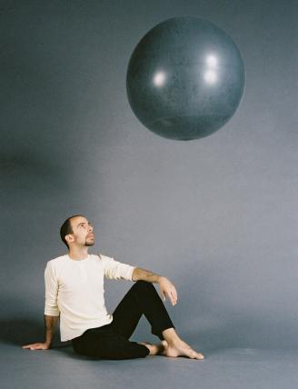 A man sits casually on the floor staring the large sphere that is floating above. 
