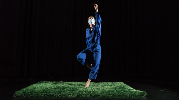 A woman in a blue jumpsuit dances on a patch of grass