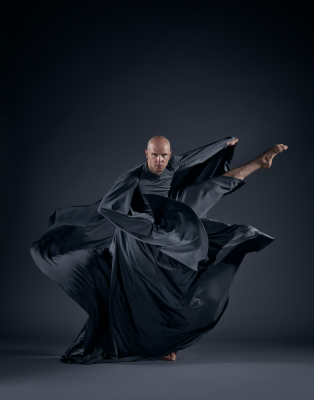 bald man in billowing dark cloak fiercely kicks his leg out to the side 