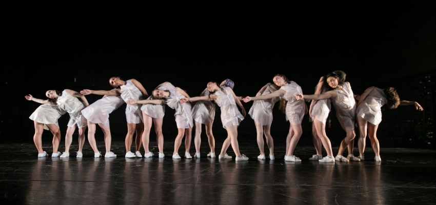 Dancers in white lean on each other in a line
