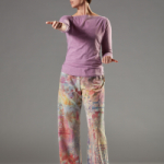Julia stands face on in colorful pants. Her right arm is extended straight forward while her left arm is bent at the elbow near her hip. Her left knee is slightly bent and she looks to her right.