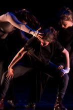 a modern dancer squats with her hands on her knees as two others reach towards her head