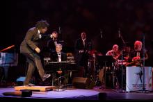 Alex Dugdale taps on stage next to a jazz band 