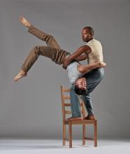 Jo stands on a chair while holding Fausto that is upside down. 