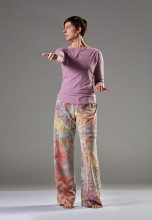 Julia stands face on in colorful pants. Her right arm is extended straight forward while her left arm is bent at the elbow near her hip. Her left knee is slightly bent and she looks to her right.