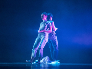 Two dancers lean back to back holding up flashlights