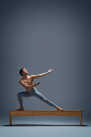 Marte lunges sideways on top of a wooden bench, with his arms and focus stretching upwards to his left. His hands are supinated, as if receiving the faint glow that is above. He is shirtless and in blue leggings. 