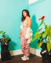 A queer mixed- Filipinx dancer poses between two plants infront of an aqua painted wall 