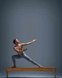 Marte lunges sideways on top of a wooden bench, with his arms and focus stretching upwards to his left. His hands are supinated, as if receiving the faint glow that is above. He is shirtless and in blue leggings. 