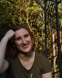 Haley smiles as they rest their forehead on their right fist. They are wearing green and there are bright trees behind them.