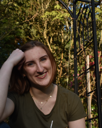 Haley smiles as they rest their forehead on their right fist. They are wearing green and there are bright trees behind them. 