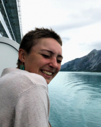 Gabbi smiles as they lean over the boat rail, with clear blue water and dark mountains behind them. 