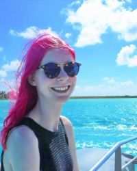 Jordan has hot pink hair and smiles with sunglasses on with the aqua blue sea behind them.  