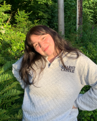 Alyssa cheerfully stands with her hands on her hips and head tilted in front of vibrant green bushes and trees. 