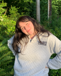 Alyssa cheerfully stands with her hands on her hips and head tilted in front of vibrant green bushes and trees.