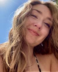 Taylor smiles as their long blonde hair falls over their bare shoulders. The sun shines brightly in the cloudless blue sky behind them. 