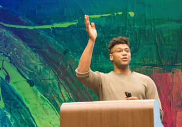 Brian J Evans giving his presentation at the 2019 Dance Graduation Research Symposium. Background is colorful painted strokes. 