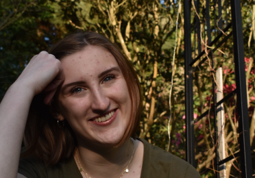 Haley smiles as they rest their forehead on their right fist. They are wearing green and there are bright trees behind them. 