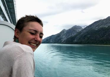 Gabbi smiles as they lean over the boat rail, with clear blue water and dark mountains behind them. 