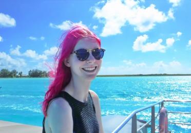 Jordan has hot pink hair and smiles with sunglasses on with the aqua blue sea behind them.  