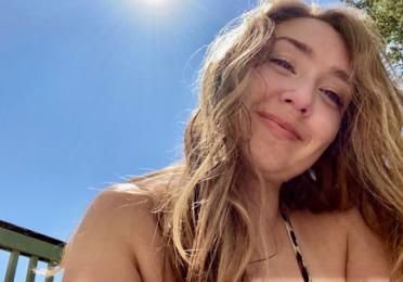 Taylor smiles as their long blonde hair falls over their bare shoulders. The sun shines brightly in the cloudless blue sky behind them. 
