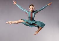 Muscular lady in striped dress jumps high with her arms out to her sides, and one foot tucked underneath. 
