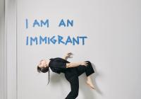 Alice dances front of a wall that says, "I am an immigrant" 