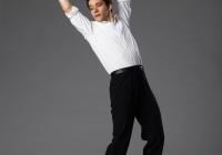 Pablo precariously balances on the front of his toes as he leans backwards,  hinging at the hips with his arms raised high above his head. He is wearing a white blouse and black pants. 
