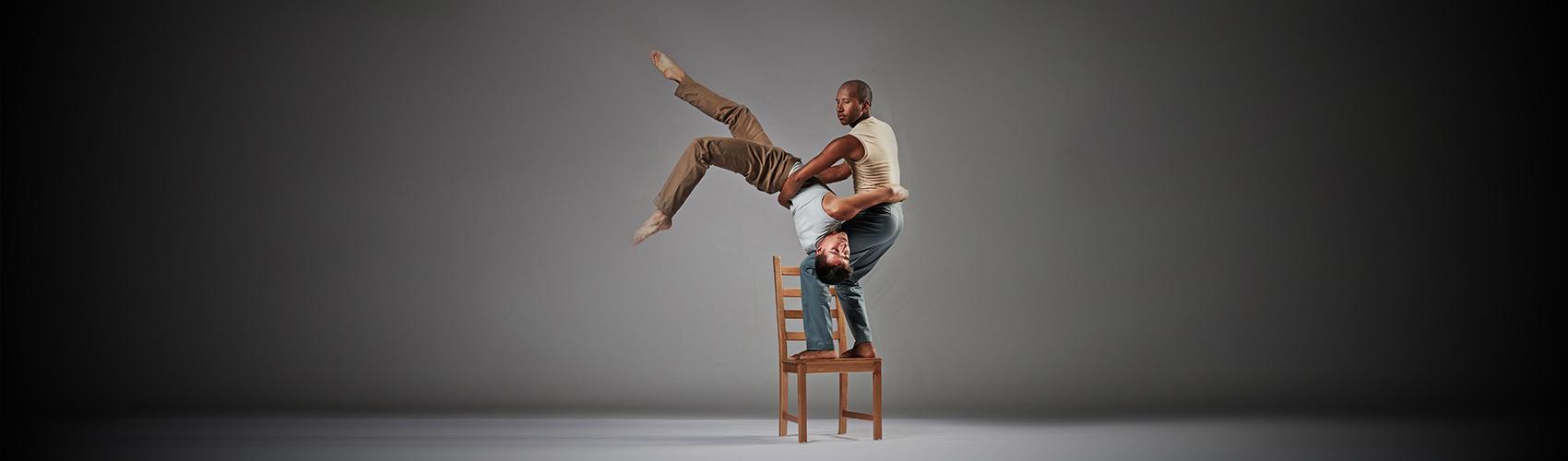 Photo of two dancers, one standing on a chair, holding up the other, who is upside-down