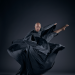 bald man in billowing dark cloak fiercely kicks his leg out to the side 