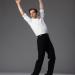 Pablo precariously balances on the front of his toes as he leans backwards,  hinging at the hips with his arms raised high above his head. He is wearing a white blouse and black pants. 